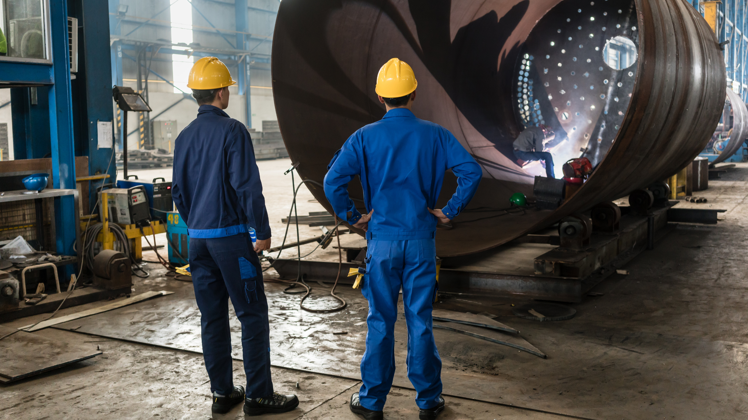 Two workers in blue uniforms and yellow hard hats observing another worker welding inside a large cylindrical metal structure in a factory setting.